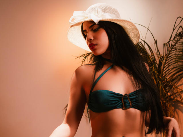 KristtieEva with long black hair, wearing a teal bikini top with a ring, and a white sun hat, her tits are small, and she has a cute, sexy look.