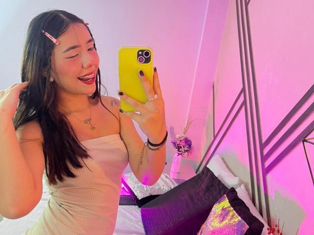 AlannaJonson with long black hair, wearing a yellow phone case, a pink hair clip, and a beige top, taking a selfie, showing her tongue out, with visible ass.