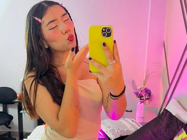 AlannaJonson with puffy lips, black nails, yellow phone, pink hairclip, white ribbed top, taking a selfie, showing off her tits in the mirror.
