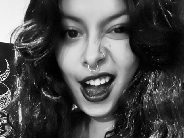 GabyJoy, con i capelli neri ricci, un anello al naso e rossetto scuro, con la bocca aperta che mostra i denti, in una foto in bianco e nero, con la lingua leggermente fuori. Le sue natiche non sono visibili..