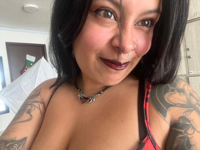 GabyJoy with black hair, nose ring, dark lipstick, red and black lingerie, tattooed arms, smiling, chest visible, necklace with red heart. Boobs in frame, medium-sized.