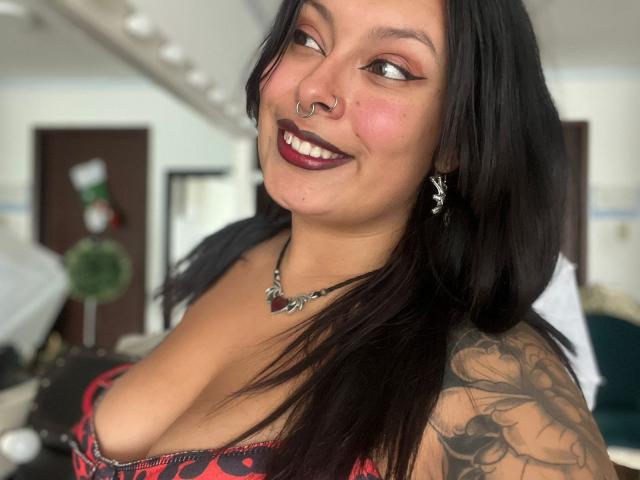 GabyJoy with dark hair, nose ring, and tattoo, smiling at camera, wearing red and black lingerie, showing her big boobs and cleavage, dark lipstick, necklace with red gem.