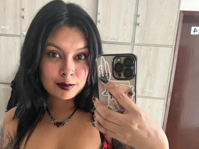 GabyJoy with dark hair, nose ring, and burgundy lipstick, taking a selfie with her phone, showing her tits and tattooed arm, wearing a red top and black necklace.