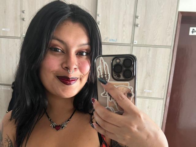 GabyJoy with black hair, nose ring, red lipstick, and tattoo, holding phone with camera, showing off cleavage in black strappy top, necklace with heart-shaped pendant.