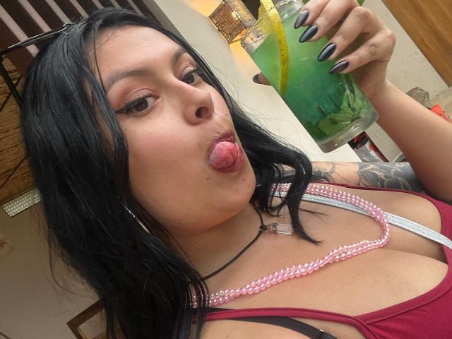 GabyJoy with black hair, pouty pink tongue out, red tank top, pink bra straps, tattoos on right arm, holding green drink with yellow straw, black nails.