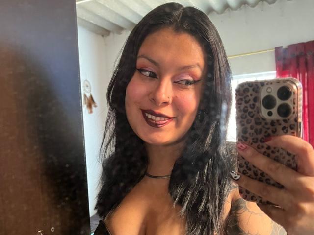 GabyJoy with dark lipstick, black hair, and pink eyeshadow, holding a leopard print phone, smiling at her reflection, showing her small tits and tattooed arm.