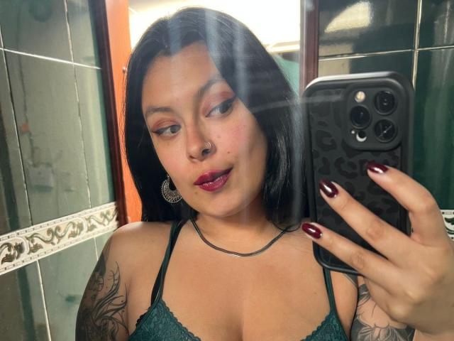 GabyJoy, con capelli scuri, ombretto rosa, rossetto rosso, piercing al naso e orecchini, che tiene in mano un telefono nero, mostra dei tatuaggi e indossa un reggiseno verde scuro, con le unghie rosse, in un selfie allo specchio..