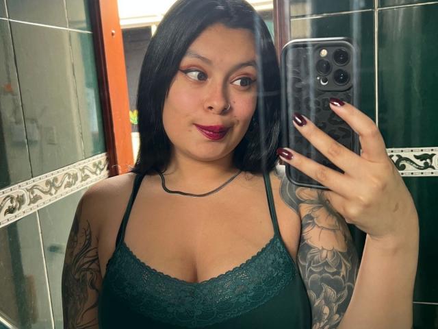 GabyJoy with dark hair, green lace bra, tattooed arm, red lipstick, holding phone, looking at mirror, showing medium tits, black nails, slight smile, green tiled bathroom.