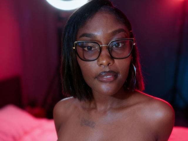 SachiZlatan222 with dark skin, short black hair, wearing glasses, and hoop earrings, looking serious with shiny lips and a tattoo on her chest. She's topless, showing her bare shoulders.