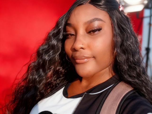 SachiZlatan222 has long, wavy black hair, brown skin, and is wearing a black and white top with a zipper. She has glossy lips and is smiling subtly.