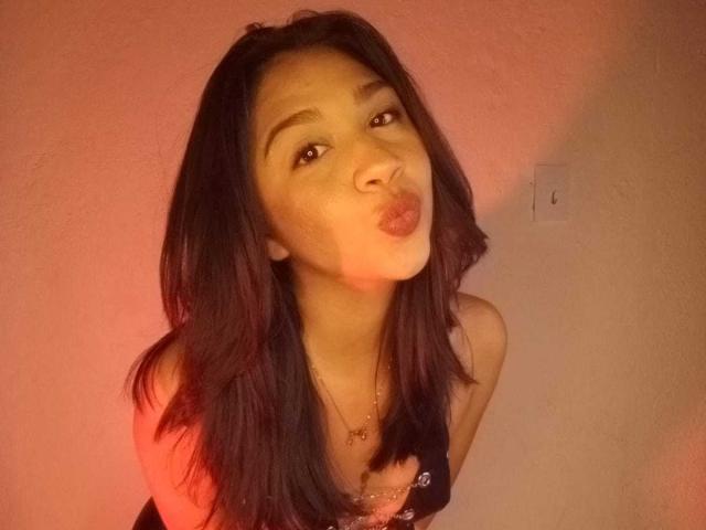 NanaMilan with dark hair, pouty lips, and a necklace, leaning forward, making a kissy face in a warmly lit room.