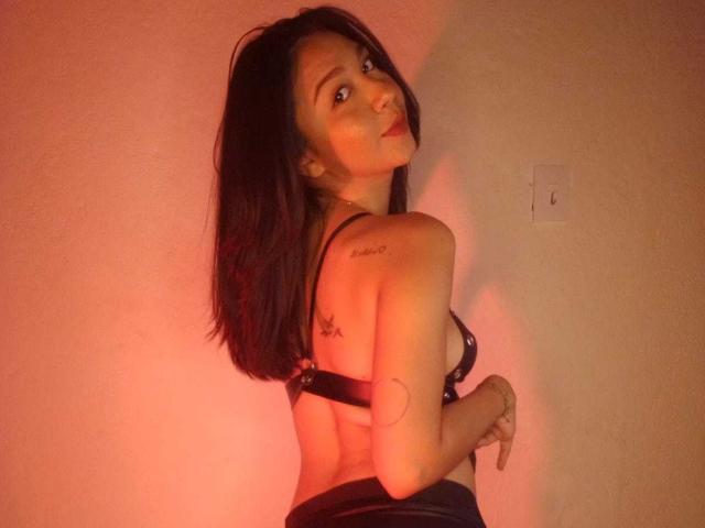 NanaMilan with dark hair, looking over her shoulder, wearing black lingerie, showing her tits, a tattoo on her right shoulder, and a ring on her left hand.