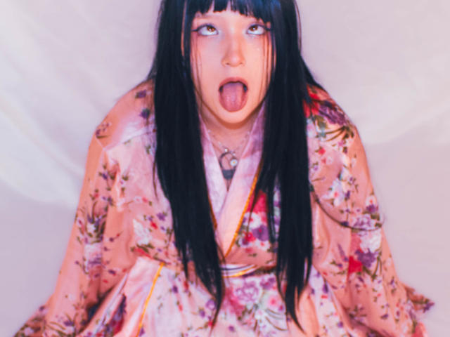 KuruChan with black straight hair, pink floral kimono, and tongue out, has a slightly flushed face, looking directly at the camera, sitting against a white background.