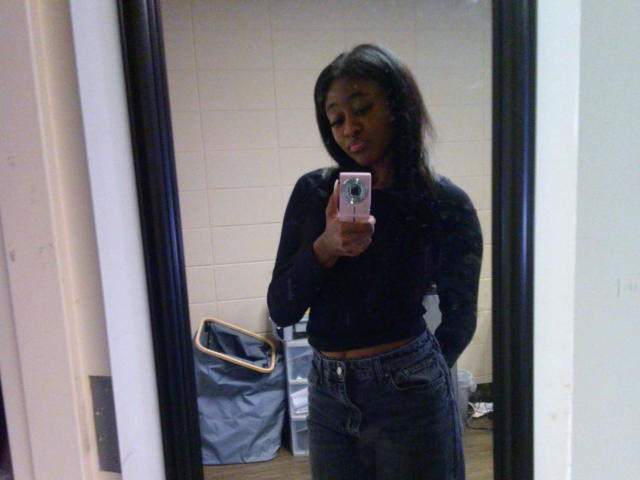 GiselleCumsForU with dark skin, long black hair, black top, and blue jeans, holding a pink phone, taking a mirror selfie, standing in a room with a bag.