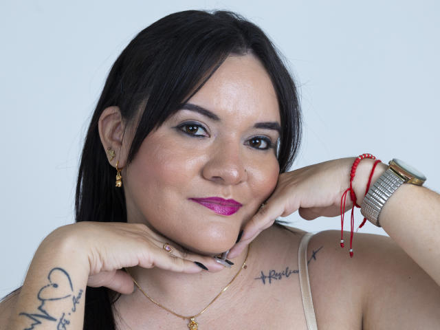 VictoriaPrice has black hair, pink lipstick, and tattoos on her arms, wearing a gold necklace and red bracelet, with hands near her face, smiling. She's got a watch too.