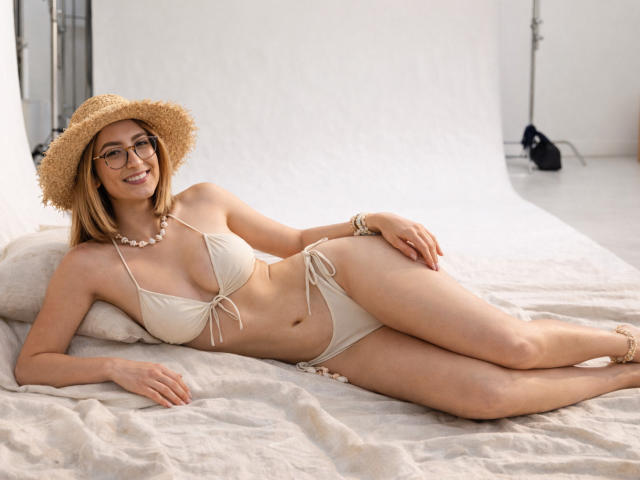 RousseBlossom with glasses, straw hat, white bikini, small tits, ass, and a pearl necklace, lying on a bed, smiling, wearing gold anklet.