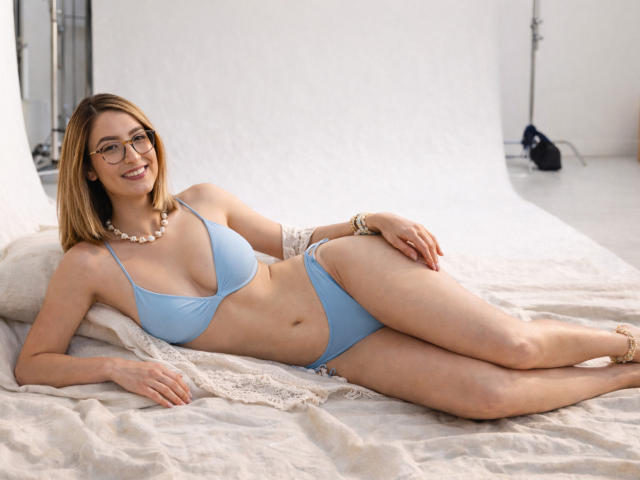 RousseBlossom, a young woman with light brown hair and glasses, lies on a white bed. She wears a light blue bikini, showcasing a slim, toned physique with small to medium-sized breasts. She has a pearl necklace and a gold bracelet. Her right arm rests on a white pillow, and her left hand is on her hip. The soft, white bedspread contrasts with her skin and blue bikini. Her relaxed, smiling pose is natural and inviting. The photograph uses soft lighting to highlight her features and the texture of the bedspread.