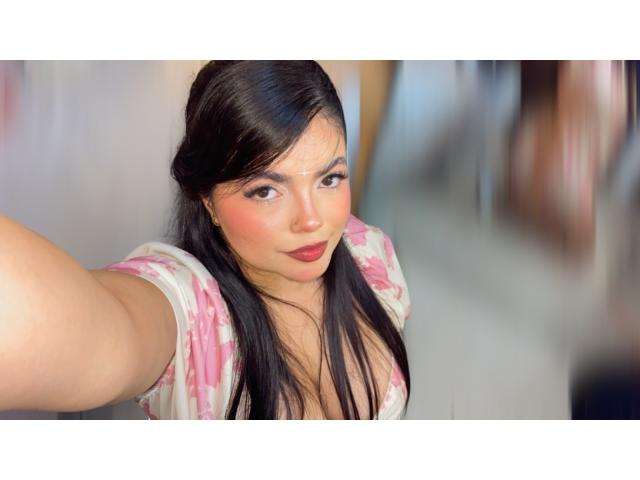 Lara James, com cabelos longos e pretos, camisa floral rosa e batom vermelho, tirando uma selfie com o braço direito, mostrando o decote e o rosto bonito, sorrindo levemente..