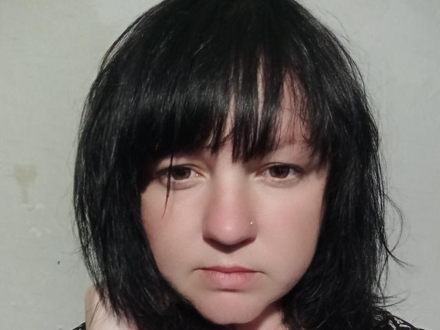 PattiLynn has black, slightly messy, medium-length hair with bangs, a pale skin tone, brown eyes, a small nose piercing, and a neutral, slightly sad expression.