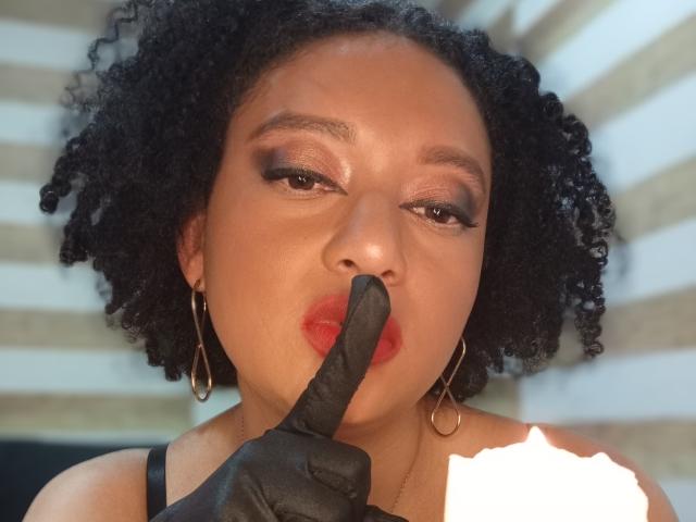VioletaSwitch with curly black hair, wearing gold hoop earrings, has red lipstick, black finger glove, and holds finger to mouth. Her brown eyes are slightly squinted.