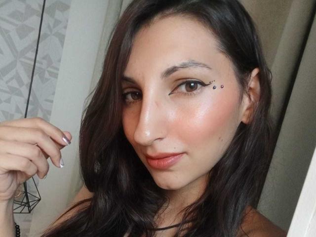 CelestePoot a les cheveux longs, bruns et ondulés, des yeux bruns, un trait d'eye-liner discret, de petits tatouages en forme de points noirs près de son œil droit, et un léger blush rose sur ses joues. Elle se tient la main sur le menton..