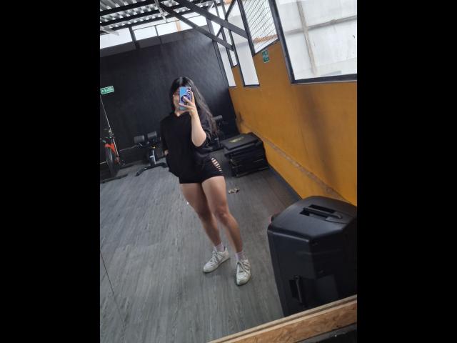 PaulethJoens in black tee with cutouts, short black shorts, and white sneakers, taking a mirror selfie, holding a blue phone, in a gym with gray floors.