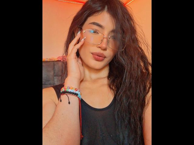 PaulethJoens met golvend donker haar, een bril en donkere lippenstift, die haar wang aanraakt, draagt een zwarte tanktop, een kleurrijke armband en steekt haar tong uit..