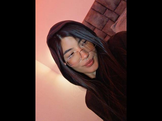 PaulethJoens, met lang zwart haar, een bril en een zwarte hoodie, glimlacht lichtjes, heeft een schattig gezicht en een rozeachtige belichting. Haar borsten zijn niet zichtbaar, alleen haar bovenlichaam..