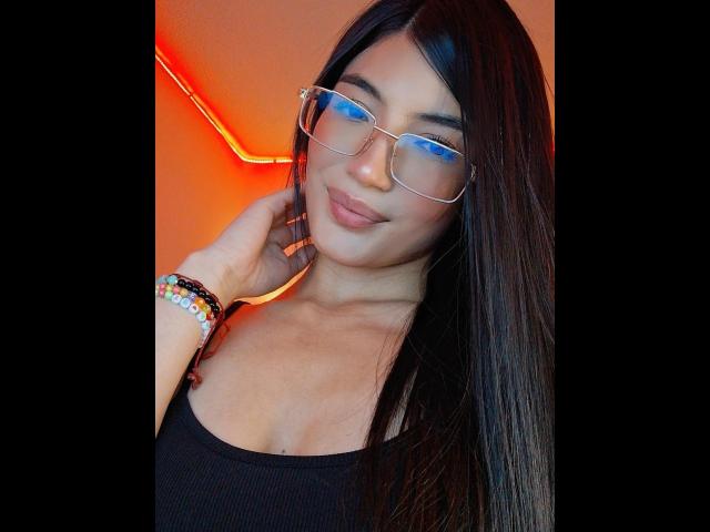 PaulethJoens with long dark hair, blue-tinted glasses, black tank top, colorful bracelets, and a subtle smile, showing her tits slightly. She's looking cute and confident.