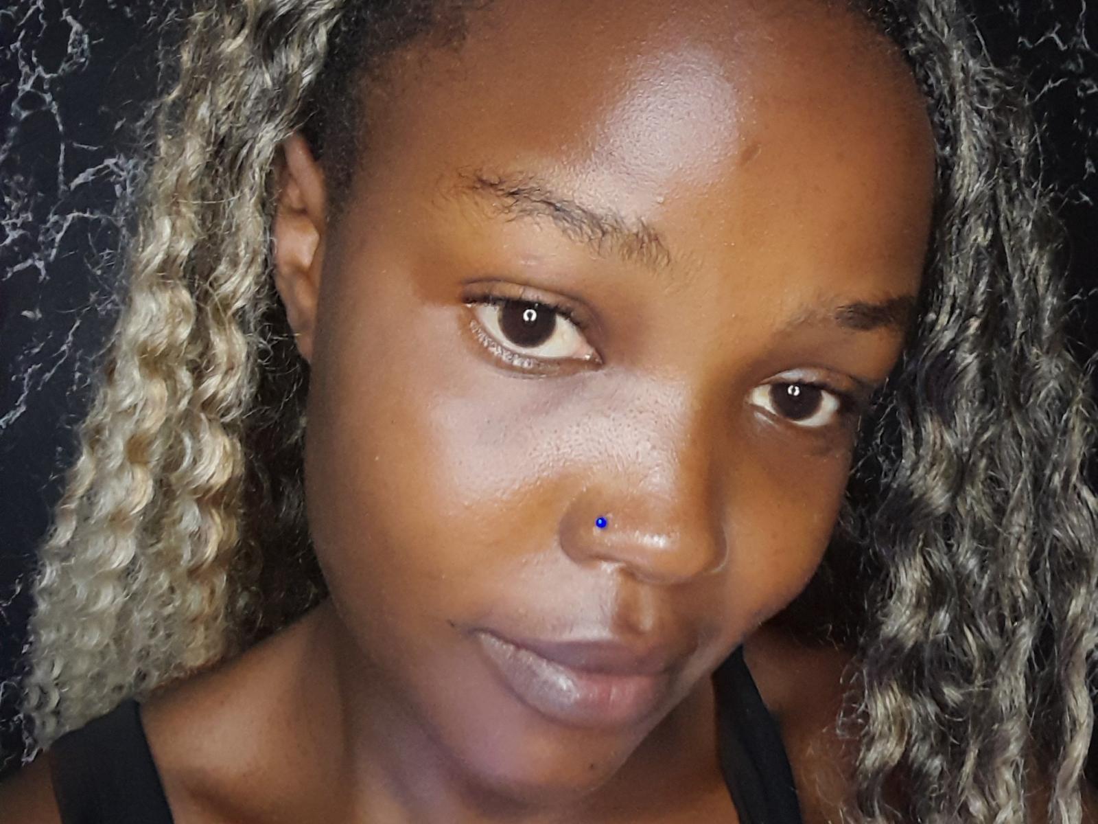 LaureneJoanna has a nose piercing with a small blue gem, curly blonde and brown hair, and a black tank top. Her skin is shiny and she's smiling softly.