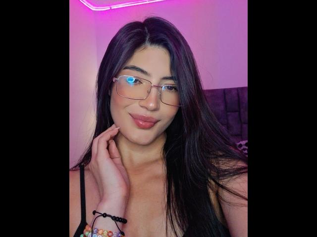PaulethJoens with long black hair, glasses, pink neon light, smiling, light skin, wearing black tank top, touching cheek, colorful bracelet on right wrist, pink lips, cute.