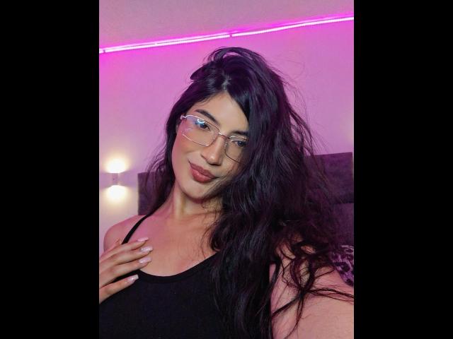 PaulethJoens with long black hair, wearing glasses and a black tank top, showing off her tits with one hand on her chest. Pink neon light in the background.