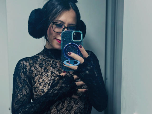 AmaraMirage with glasses, black lace top, and double buns, holding a phone covering her tits, red lipstick, and dark nails, standing in a mirror selfie.
