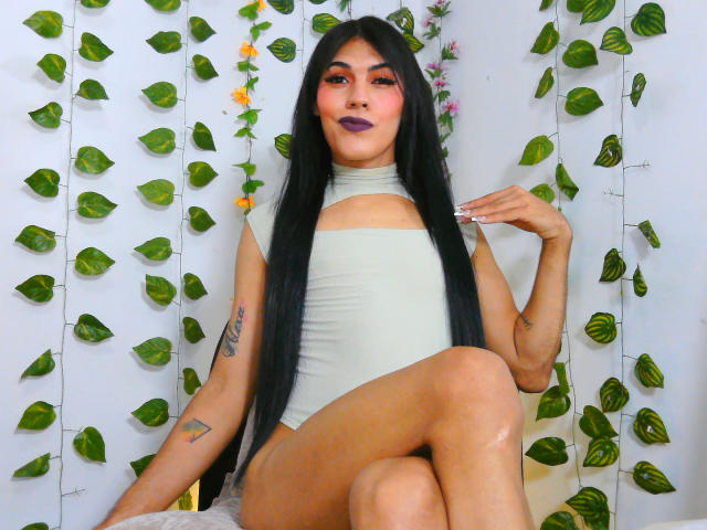 AshleyCock with long black hair, wearing a green cut-out top, purple lipstick, and tattoos, sitting with legs crossed, showing off her smooth, tan skin and small ass.