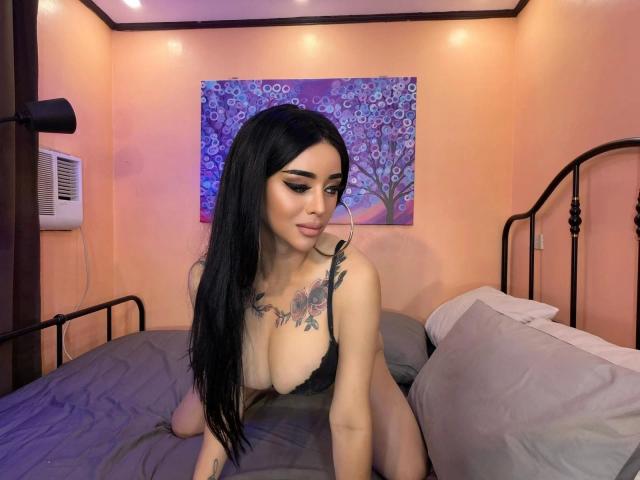 GoyaLakinova-hot is a young woman with long black hair and fair skin, sitting on a bed with gray sheets. She has large, exposed breasts and a floral tattoo across her chest. She wears a black lace bra, with her arms resting on the bed, and looks down with a slight smile. The room has peach-colored walls and a purple floral painting in the background. The bed has a black metal frame and a white pillow. The lighting is soft and warm.