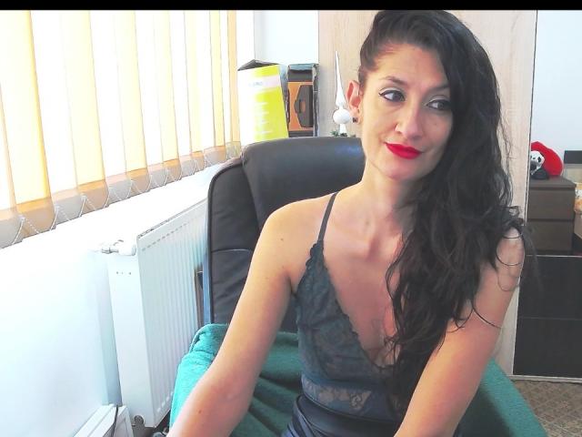 BeatrixCharmm with long black hair, red lipstick, and a deep cleavage in a black lace top, sitting in a chair, green towel on her lap, smiling.