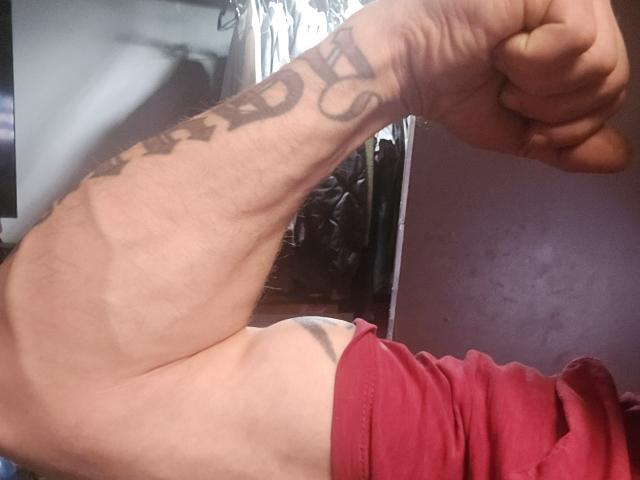 ShyGuyy's arm is flexed, showing a tattoo with bold letters. His red shirt is pulled up, exposing a sliver of his white underwear. His hand is clenched.