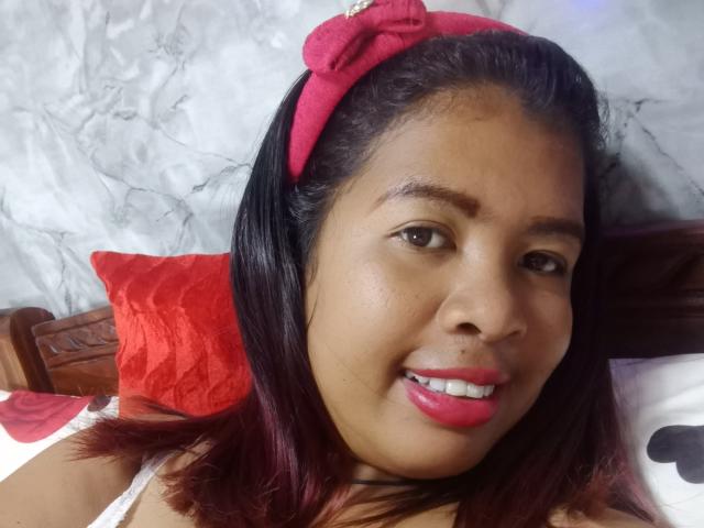 MariahSquirting69, com pele morena, faixa vermelha na cabeça, batom rosa, sorrindo, cabelo preto, travesseiro vermelho, cama branca e vermelha, parede de mármore, olhando para a câmera, plano médio, parte superior do corpo..