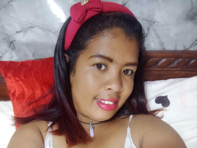 MariahSquirting69 with dark brown skin, red lipstick, and pink bow in black hair, wearing a white lace top and purple necklace, smiling at the camera.