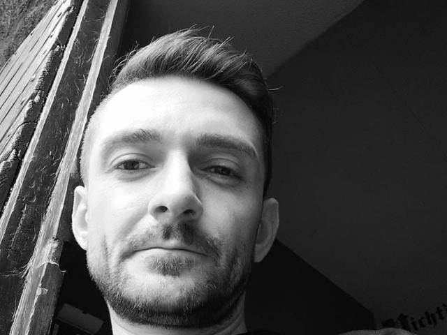 Black-and-white selfie of Unterwegsmitmir, a bearded dude with a slight smirk, short dark hair, and a clean-shaven face, leaning against a rough wooden surface.