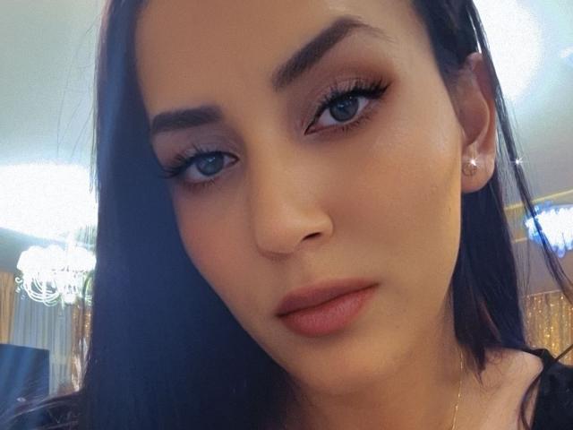 WildFoxxy69 has blue eyes, full pink lips, and dark brown hair, wearing pink earrings, with subtle makeup, looking directly at the camera with a slight, sexy, confident smirk.