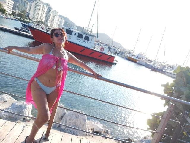 Yeseniaa69 with pink cover-up, white bikini bottom, and pink bikini top, leaning on metal railing, water and boats in background, wearing sunglasses, standing on wooden dock.