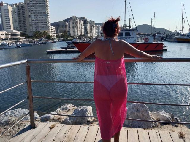 Yeseniaa69 in a pink sheer dress, back to camera, showing her ass, stands on a wooden dock, arms on a metal railing, overlooking a marina with boats.