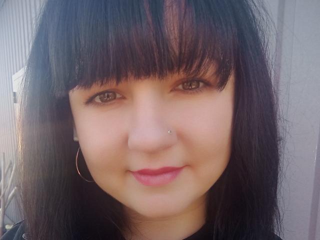 PattiLynn has black straight hair, brown eyes, a nose piercing, and a subtle smile with pink lipstick. She wears a black top and a small hoop earring.