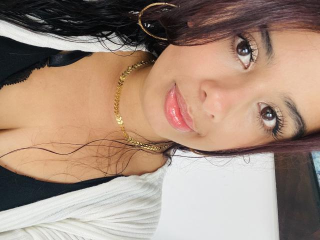 KeyklinOne with shiny pink lips, brown eyes, and wavy dark hair, wearing a gold chain necklace, hoop earrings, black top, and white cardigan, smiling at the camera.