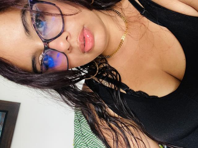 KeyklinOne tiene gafas azules reflectantes, labios rosados y brillantes, cabello negro ondulado, un collar dorado, un sujetador negro y un busto curvilíneo con escote visible. Su piel es morena..