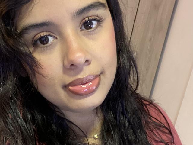 KeyklinOne has shiny brown eyes, dark wavy hair, and glossy pink lips. Her tan skin is smooth, and she's wearing a pink shirt with a gold necklace.