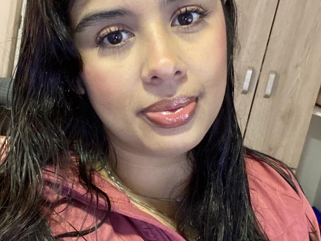 KeyklinOne's face, shiny pink lips, dark wet hair, brown eyes, slight smile, pink shirt, light brown skin, wooden cabinet in background, subtle makeup, glossy lips.