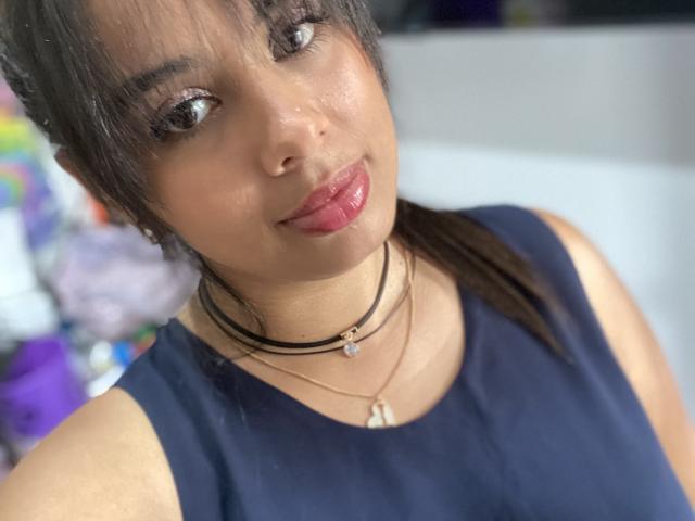KeyklinOne with shiny brown eyes, glossy pink lips, and dark hair in a ponytail, wearing a blue top, black choker, and necklace, smiling slightly. Her skin is tan.