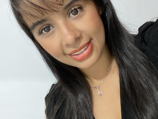 KeyklinOne ha la pelle marrone media, lunghi capelli neri con frangia, occhi marroni e rossetto rosso. Sta sorridendo, indossa una maglietta nera e una collana d'argento con una piccola croce..